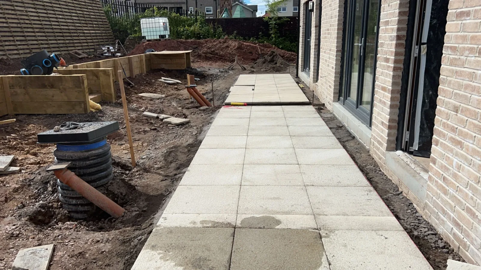 Porcelain slab path running along the side of a new-build home, with raised sleeper beds and a drainage inspection chamber under construction alongside.