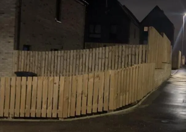 New pale timber fence running along a wet street and pavement at night under a streetlamp.