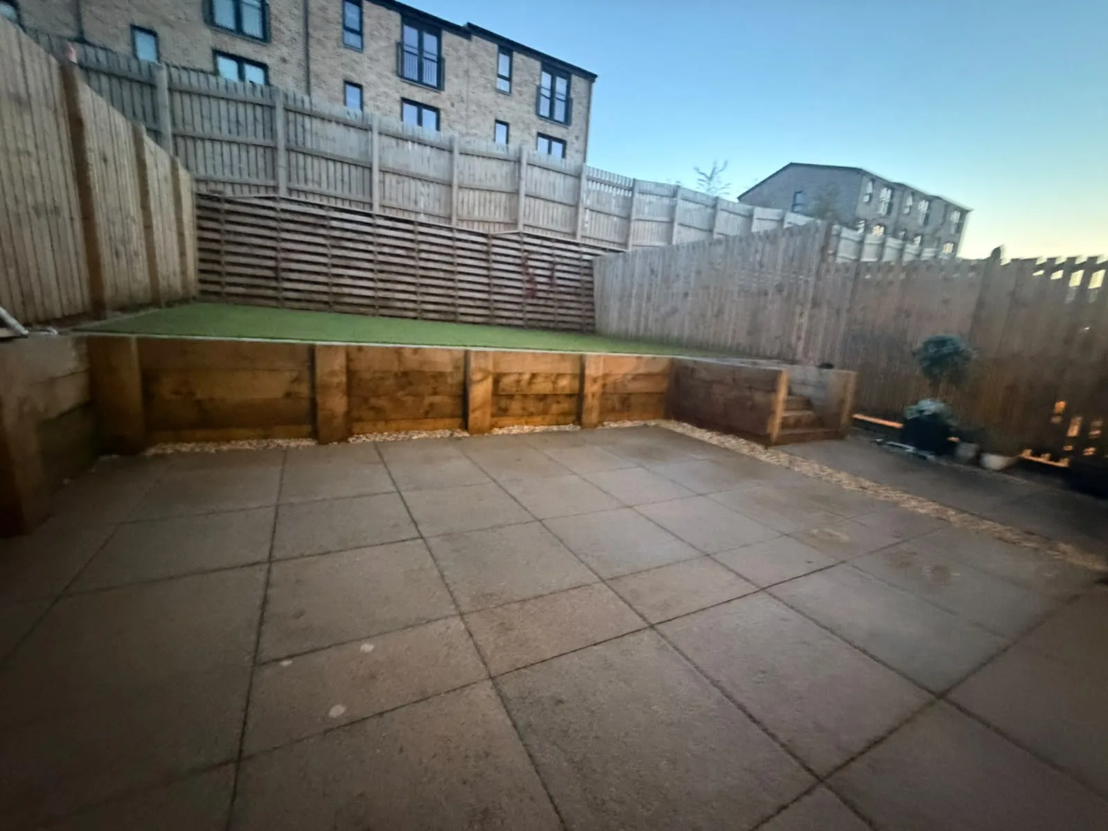 Porcelain patio at dusk with raised sleeper walls, artificial turf and timber fencing.