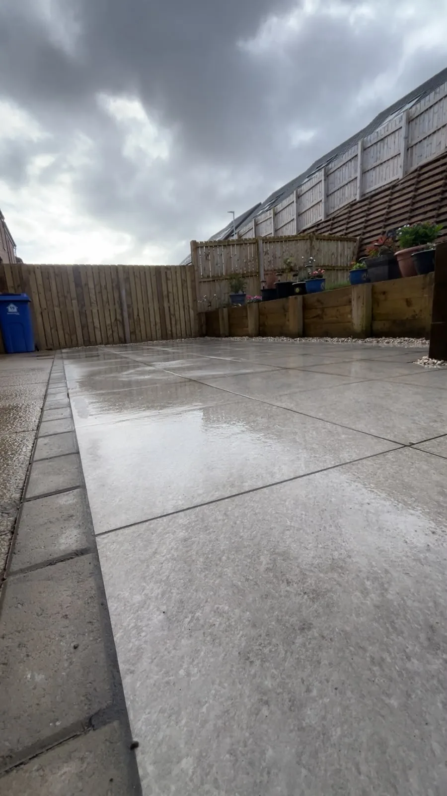 New grey porcelain patio with a monoblock paving border on a wet day, beside sleeper raised beds and timber fencing.